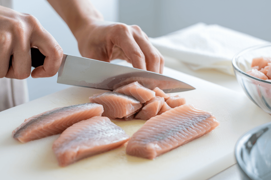 A close-up of a cutting board with boneless, skinless fish fillets being prepared. The scene shows clean hands using a knife to cut the fillets into small chunks.