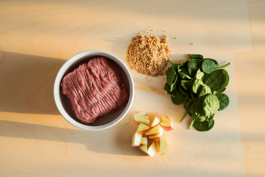 Image of a bowl of raw, ground turkey, a small pile of cooked quinoa, a handful of fresh spinach leaves, and a few finely chopped apple cubes.