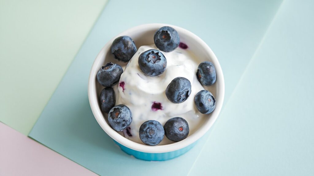 A playful image of frozen yogurt and blueberry bites inside a small, white bowl set on a light pastel surface.