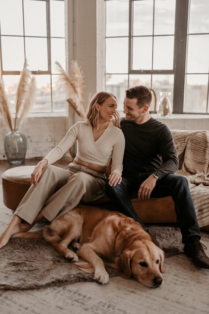 Editorial-style at-home engagement shoot a couple sits on a modern bench in front of floor-to-ceiling windows, gently petting their golden retriever lying at their feet