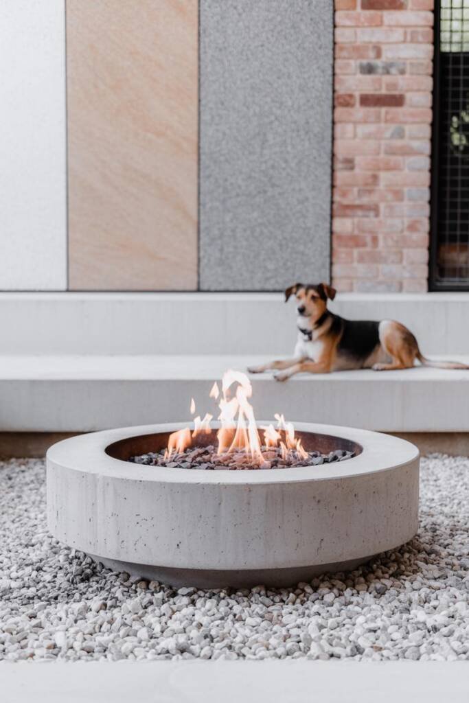 Vertical 2:3, close-to-mid shot of a concrete/stone fire pit ring on crushed gravel, flames low; swatch-style composition sho